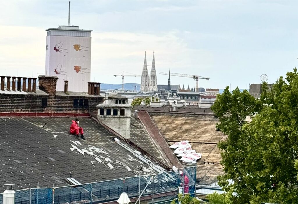 Arbeiter in roten Schutzanzügen und mit FFB2-Masken entfernen asbesthaltige Eternitplatten vom Dach der Schule Schwarzingergasse